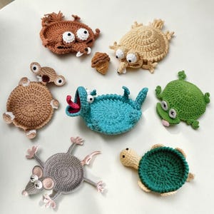 May include: A collection of handmade crochet animal coasters in various colors. The set includes a brown creature with large white eyes, a light brown creature, a blue creature with a red tongue, a green frog, a gray mouse, and a green and tan turtle.