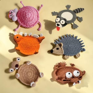 Könnte beinhalten: Eine Sammlung von sechs handgefertigten Tieruntersetzern aus Häkelarbeit. Die Untersetzer umfassen ein rosa Wesen, einen grauen Waschbären, einen orangefarbenen Dinosaurier, einen blauen Igel, eine beige Schildkröte und ein braunes Wesen mit großen Augen.
