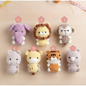 Może przedstawiać: Kolekcja siedmiu ręcznie robionych zabawek zwierzęcych na szydełku, w tym słoń, lew, małpa, niedźwiedź, żyrafa, tygrys i kolejny niedźwiedź. Każda zabawka ma inny kolor, szczegółowe cechy i miękką fakturę.