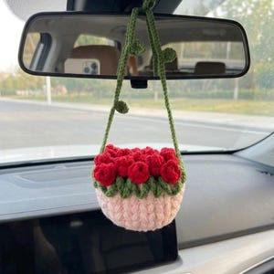 May include: A handmade crochet car charm featuring a small basket filled with red roses. The basket is pink and green, with a green stem and leaves. The charm hangs from a car's rearview mirror.