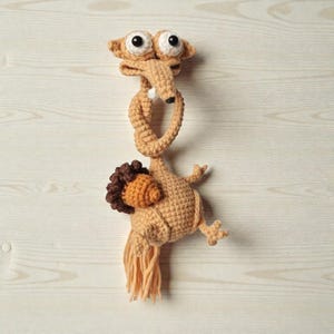 Peut inclure: Une créature au crochet faite à la main, dans des tons de beige et de marron, avec de grands yeux expressifs et un cou noué. La créature a une touffe de cheveux bruns et une frange de fil beige. Le design fantaisiste est posé sur un fond clair en bois.