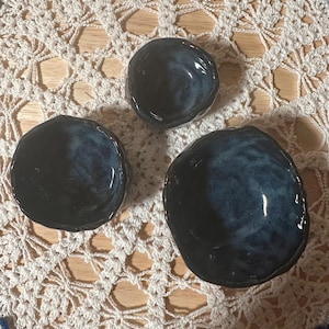 May include: Three small, handmade ceramic bowls with a dark blue glaze. The bowls have irregular, organic shapes and sit on a white crocheted doily. The glaze has a glossy finish, and the bowls vary slightly in size.