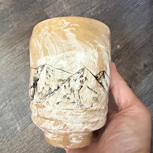 May include: A cylindrical ceramic cup with a textured, light tan clay body. The exterior features a hand-painted white wash overlay and detailed black ink illustrations of mountain peaks and small birds flying in the sky above the range.