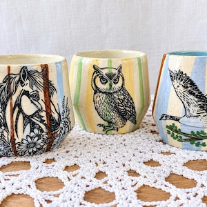 May include: Three ceramic cups featuring hand-drawn illustrations. The first cup shows a horse with flowers, the second an owl, and the third a goose in flight. Each cup has a cream-colored base with vertical pastel blue, yellow, and brown stripes.