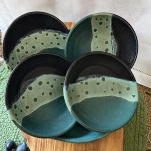 Può includere: Un set di quattro ciotole rotonde in ceramica con esterno nero scuro e smalto interno bicolore. Le superfici interne sfumano da una base color verde acqua profondo a una fascia verde salvia più chiara decorata con macchie circolari scure.