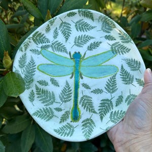 May include: A round ceramic plate with a white base featuring a central light blue dragonfly illustration. The dragonfly has a dark blue body and is surrounded by numerous small, green fern leaf imprints scattered across the entire surface.