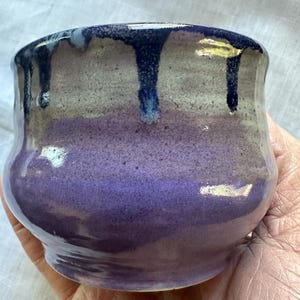 Puede incluir: Un cuenco de cerámica hecho a mano con forma redondeada y superficie texturizada. El cuenco presenta un degradado de colores, pasando de un morado intenso en la base a un gris más claro en la parte superior, con gotas azul oscuro en el borde.