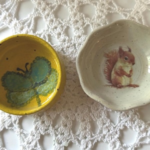 Op de afbeelding: Twee kleine keramische schaaltjes met een gespikkelde afwerking. Het linker schaaltje is felgeel en voorzien van een geschilderde groene vlinder. Het rechter schaaltje is gebroken wit met een geschulpte rand en een afbeelding van een eekhoorn.