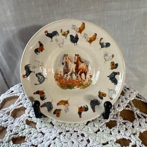 May include: A round ceramic plate with a speckled cream finish featuring a central illustration of two horses running through orange flowers, surrounded by a border of various roosters and hens in shades of black, brown, white, and red.