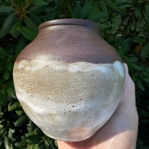 May include: A small, rounded ceramic vase featuring a smooth, dark brown upper section that transitions into a textured, speckled glaze. The lower portion displays a gradient of light beige and white tones with a porous, cratered surface finish.