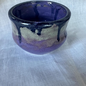 May include: A handmade ceramic bowl with a rounded shape. The bowl features a dark purple rim with dripped glaze over a light grey and purple body. The interior is a deep purple color. The bowl is likely used for serving or display.