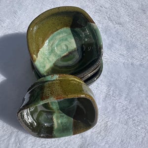 May include: A collection of handmade ceramic bowls. Each bowl has a unique colour scheme with shades of green, brown, and black. The bowls have a glossy finish and a square shape with rounded edges. The bowls are stacked on top of each other.