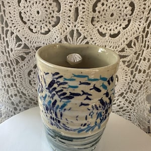May include: A cylindrical ceramic vessel with a speckled off-white glaze, featuring a hand-painted pattern of small dark blue and light blue fish swimming in a school. The upper rim has a small circular cutout, and the base has horizontal ridges.