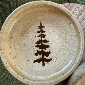 May include: A handmade ceramic bowl with a speckled, beige glaze. A brown tree design is painted in the center. The bowl has a slightly raised rim and is suitable for serving food. The bowl is approximately 8 inches in diameter.