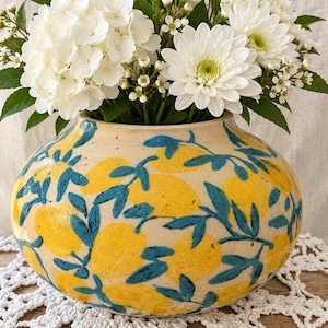 May include: A round ceramic vase with a cream-colored base featuring a hand-painted pattern of bright yellow lemons and dark teal green leaves. The vase is filled with a large bouquet of white hydrangea and daisy flowers with green foliage.