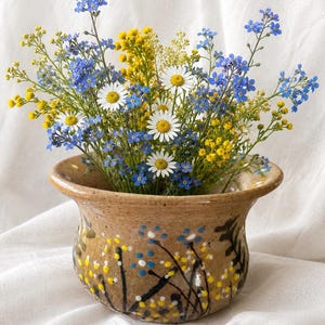 May include: A tan ceramic planter with a flared rim, decorated with hand-painted blue, yellow, and white floral patterns. The planter is filled with a bouquet of fresh blue forget-me-nots, yellow mimosa, and white daisies with yellow centers.
