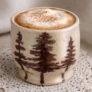Può includere: Una tazza di ceramica con un disegno di foresta, piena di cappuccino. La tazza ha una base color crema con illustrazioni di alberi marroni. Il cappuccino ha uno spesso strato di schiuma, cosparso di spezie.