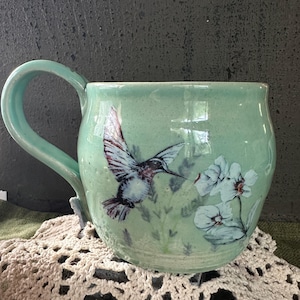 May include: A white ceramic mug with a black handle and the text "But First, Coffee" in a black, bold font. The mug is on a white surface. The mug is approximately 4 inches tall.