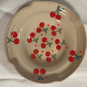 May include: A beige ceramic plate with a scalloped edge, adorned with hand-painted red cherries and green stems. Small red hearts are scattered around the cherries. The plate has a slightly speckled texture.