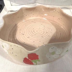 Puede incluir: Un cuenco de cerámica beige con borde ondulado. El cuenco presenta motas oscuras y diseños florales pintados a mano en rojo, blanco y verde. El interior tiene un patrón en espiral.