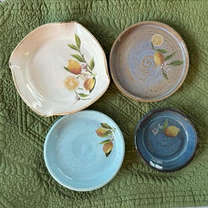 May include: Four ceramic plates with lemon designs. The plates are in various shapes and colors, including white, blue, and brown. Each plate features hand-painted lemons and green leaves. The plates are arranged on a green quilted surface.