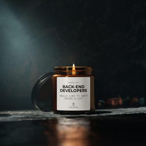 May include: A lit candle in a brown glass jar with a gold lid. The label reads "CANDLE FOR BACK-END DEVELOPERS" and "SMELLS LIKE 'I CAN'T CENTER A DIV'". The candle is on a dark surface with a dark background.