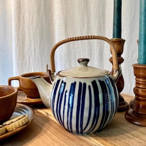 May include: A ceramic teapot with a cream-colored body and blue vertical stripes. It has a woven handle and a matching lid. The teapot is part of a tea set, with a wooden teacup and saucer visible.