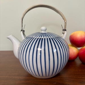May include: A white ceramic teapot with a rounded body and a lid, adorned with vertical blue stripes. It features a woven handle and a spout. Apples are in the background. The teapot is likely for brewing tea.