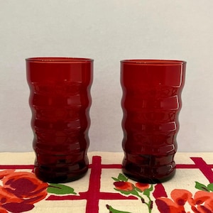 May include: Two ruby red drinking glasses with a textured, wavy design. The glasses are cylindrical and sit on a floral patterned tablecloth. The glasses are made of a transparent material.