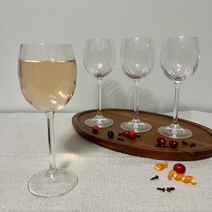 May include: Four clear wine glasses, one filled with a light pink liquid, sit on a wooden oval tray. The other three glasses are empty. Garnishes of red berries, cloves, and orange zest are scattered around the tray.