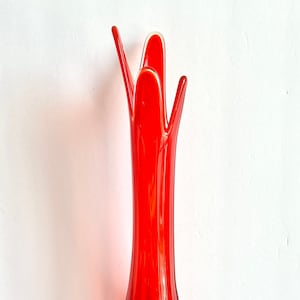 May include: A tall, vibrant red glass vase with a flared top and a bulbous base. The vase has a unique, elongated shape and sits on a yellow-orange, petal-like base. The glass appears to be transparent.