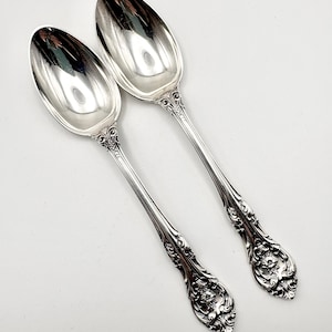 May include: Two polished silver-toned spoons featuring intricate embossed floral and scrollwork detailing on the handles. The spoons have smooth, reflective oval bowls and ornate, decorative stems that taper toward the base of the handle.