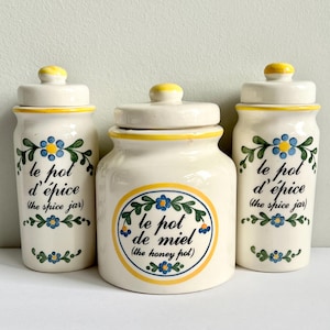 May include: Three off-white ceramic spice jars with yellow accents and floral designs. The jars are labeled "le pot d'épice" and "le pot de miel" in French. The jars have lids with yellow knobs.