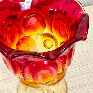 May include: A vintage red and yellow glass bowl with a scalloped rim. The bowl features a starburst pattern on the base and interior. The glass has a vibrant, translucent quality, with a gradient of colors from red to yellow.