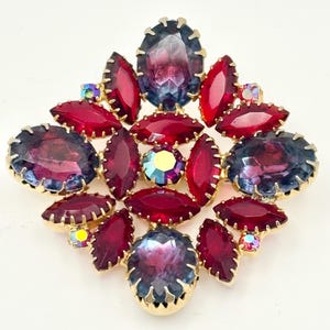 Op de afbeelding: Een vintage broche met een goudkleurige zetting, met grote, gefacetteerde edelstenen in tinten rood, paars en blauw. De broche heeft een ingewikkeld ontwerp met verschillende vormen en maten stenen, wat een schitterend effect creëert.