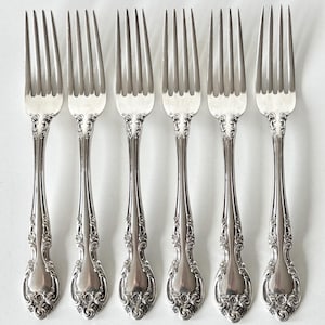 May include: Six silver forks with ornate floral and scroll detailing on the handles. The tines are evenly spaced and the forks are arranged in a row against a white background. The silverware has a classic, elegant design.
