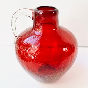Puede incluir: Una jarra de vidrio rojo vibrante con un asa transparente. La jarra tiene un cuerpo redondo y una superficie texturizada y agrietada. El asa está unida a la parte superior. Ideal para servir bebidas, con una capacidad de aproximadamente 1,5 litros.