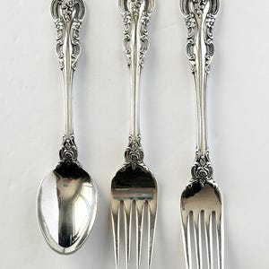 May include: A set of three silver-coloured metal utensils consisting of one spoon and two forks. Each piece features an ornate, raised floral and scrollwork pattern on the handle and the neck, with a polished, reflective finish on the bowl and tines.