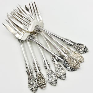Puede incluir: Un conjunto de tenedores plateados ornamentados, dispuestos sobre una superficie blanca. Los tenedores tienen intrincados diseños florales en los mangos y las púas. Probablemente para cenas formales.