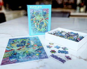 May include: A 500-piece jigsaw puzzle with an abstract art design in shades of blue, green, and red. The puzzle is partially assembled, with pieces scattered near the box and the completed image. The box is light blue.