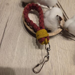 Könnte beinhalten: Ein handgefertigter Schlüsselanhänger mit einem rot-gelben geflochtenen Design. Der Schlüsselanhänger hat einen silberfarbenen Verschluss zum Befestigen an Schlüsseln oder Taschen. Der Hintergrund umfasst Baumwollblüten und eine Holzoberfläche.