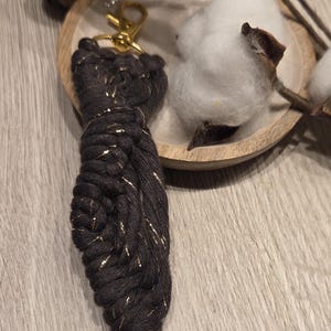 Puede incluir: Un llavero de macramé marrón oscuro con detalles metálicos dorados. El llavero está anudado con un patrón decorativo y tiene un cierre dorado. El fondo incluye motas de algodón y un plato de madera.