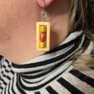 Könnte beinhalten: Ein rechteckiger, cremefarbener Ohrring mit einem silbernen Haken. Der Ohrring hat ein vertikales Design mit zwei gelben Perlen und einer orangefarbenen Perle im Rahmen. Der Ohrring wird am Ohr getragen.