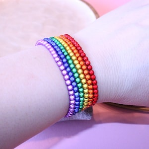 Könnte beinhalten: Ein regenbogenfarbenes Perlenarmband. Das Armband besteht aus kleinen, runden Perlen in einem Farbspektrum, darunter Rot, Orange, Gelb, Grün, Blau und Lila. Das Armband wird am Handgelenk getragen.