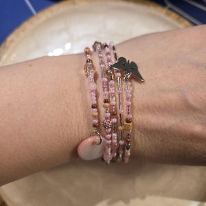Può includere: Un insieme di braccialetti di perline nei toni del rosa, marrone e argento. Un braccialetto presenta un ciondolo a farfalla, e un altro ha un ciondolo ovale rosa. I braccialetti sono impilati su un polso, mostrando la delicata lavorazione.