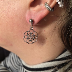 Könnte beinhalten: Silberner Ohrring mit kleinem Kugelstecker und einem hängenden, durchbrochenen Design aus ineinandergreifenden Kreisen. Der Ohrring wird an einem Ohr mit mehreren Piercings getragen. Minimalistischer Stil.