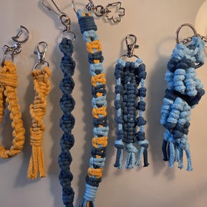 Könnte beinhalten: Eine Sammlung handgefertigter Makramee-Schlüsselanhänger in verschiedenen Designs und Farben. Die Schlüsselanhänger weisen komplizierte Knüpfmuster in Gelb-, Blau- und Mischfarben auf. Jeder Schlüsselanhänger ist mit einem silberfarbenen Metallverschluss versehen.