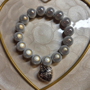 Könnte beinhalten: Ein graues Perlenarmband mit einem silbernen Herzanhänger. Das Armband hat große, runde Perlen mit einem perlmuttartigen Glanz. Der Herzanhänger ist aufwendig mit einem floralen Design verziert. Das Armband wird auf einem herzförmigen Goldrahmen präsentiert.