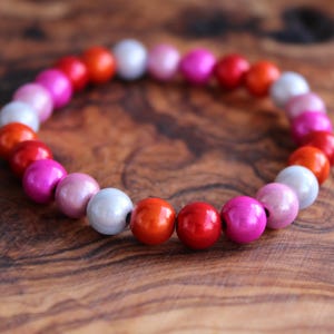 Könnte beinhalten: Ein Armband mit bunten Perlen in Rot, Orange, Rosa und Weiß. Das Armband liegt auf einer Holzoberfläche. Die Perlen haben einen Durchmesser von etwa 0,8 cm.