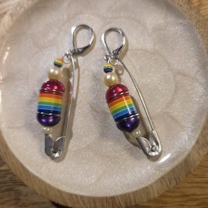 Könnte beinhalten: Ein Paar handgefertigte Ohrringe mit einem zylindrischen Perlendesign in Regenbogenfarben, Akzenten aus Perlen und einer silbernen Sicherheitsnadel. Die Ohrringe haben einen Klappbügelverschluss.